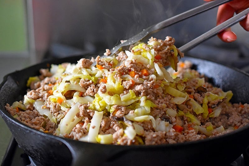 Tossing the ingredients for egg roll in a bowl with a pair of metal tongs