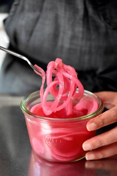 Someone in a dark grey apron is using a fork to pull some quick pickled red onions out of a jar.