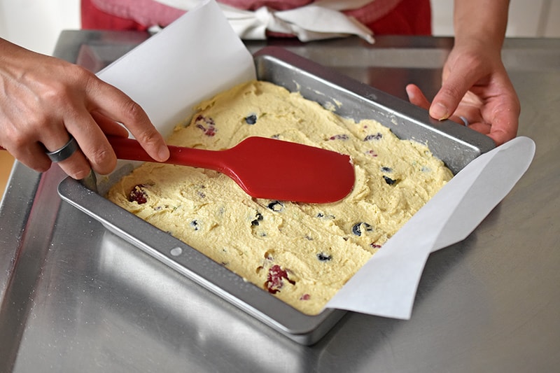 A red silicone spatula is smoothing the top of berry snack cake batter in a parchment lined square metal pan.