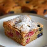 A slice of paleo and gluten-free berry snack cake is on a white plate. In the background are the other slices of berry snack cake.