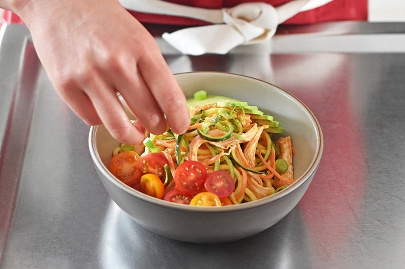 A bowl of spicy Thai chicken zoodle salad topped with tomatoes.