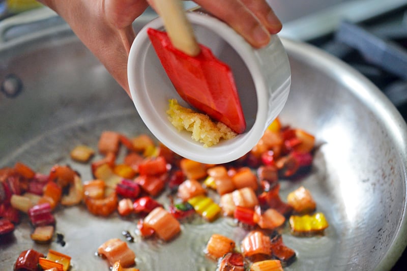 Adding minced garlic to a pan of sautéed swiss chard stems.