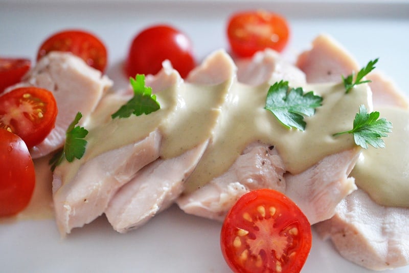 A white plate topped with poached chicken and sliced cherry tomatoes, drizzled with homemade Tonnato Sauce 