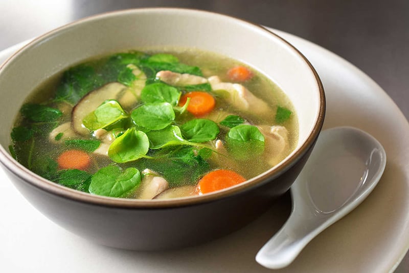 A side view of a bowl of Watercress & Chicken Soup on a plate with a white ceramic spoon next to it.