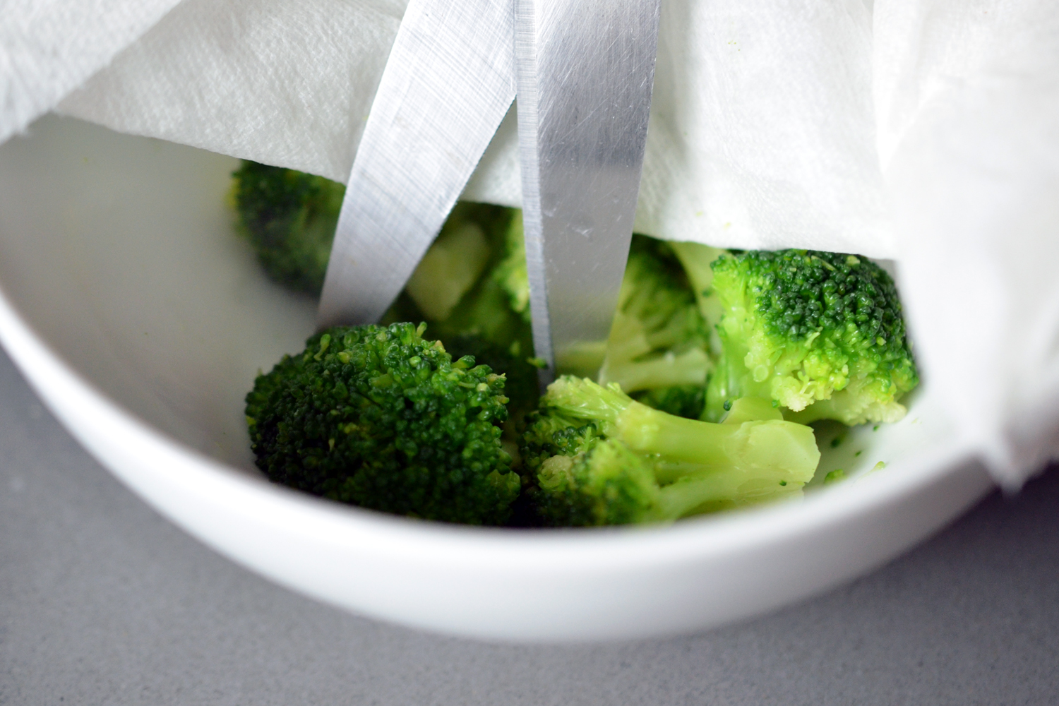 A close up side view of a bowl of microwaved frozen broccoli. The broccoli is being cut up with a pair of kitchen shears.