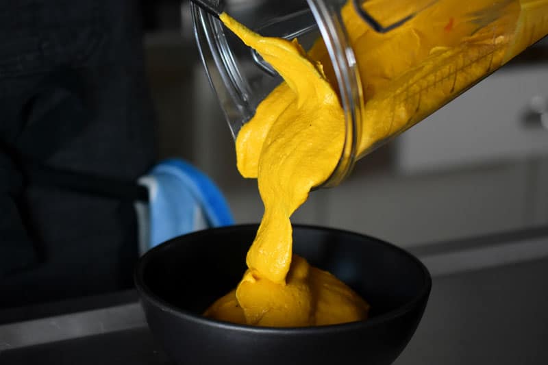 Someone pouring the bright orange-yellow sweet potato and cashew dip into a bowl.