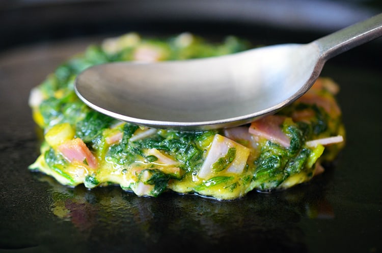 An Egg Foo Young (Spinach, Egg, Ham & Coconut Pancakes) is being fried on a cast iron skillet.