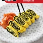 A white plate with a gray flower pattern is topped with a Korean rolled egg omelet with seaweed. There is a small pile of kimchi on the side and a pair of chopsticks is lifting up a piece. The red banner at the bottom says paleo, keto, vegetarian, and gluten-free.
