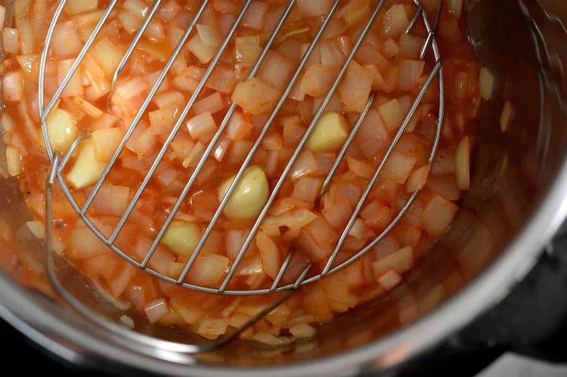 A stainless steel steaming rack is placed on top of the cooked alliums in an Instant Pot.
