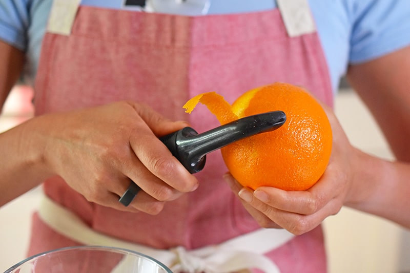 A person in a red apron is peeling the zest off of an orange with a vegetable peeler.