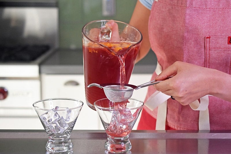 Pouring a pomegranate orange juice mocktail through a strainer and into a fancy glass for serving.