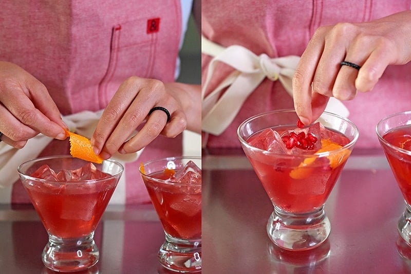A person in a red apron is adding a piece of twisted orange zest and pomegranate arils to a glass filled with a pomegranate orange juice mocktail.