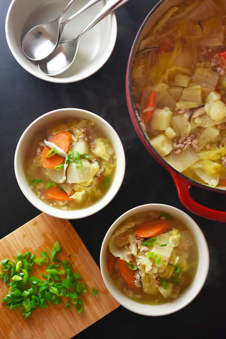 An overhead shot of a tabletop with bowls of Pork and Napa Cabbage Soup and a cutting board topped with sliced scallions