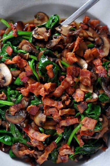 An overhead shot of a plate filled with sautéed spinach, crispy bacon bits, shallots, and mushrooms.