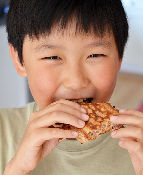 A smiling Asian boy is eating a Bacon Pancake Sandwich.