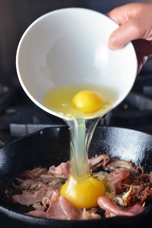 A hand is pouring two eggs into a skillet filled with prosciutto and kalua pig to make Pig in a Skillet.