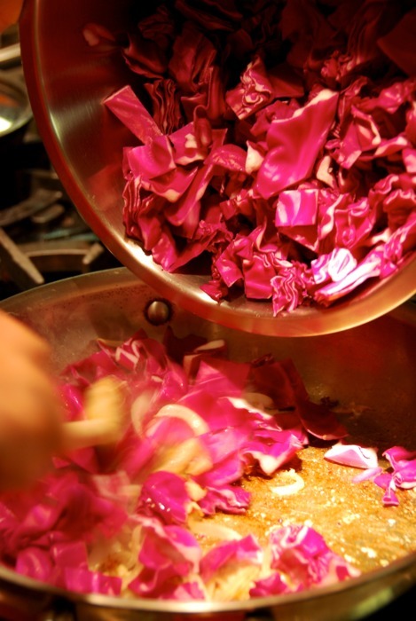 Someone adding a large bowl of red cabbage to a stainless steel pan.