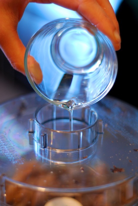 A hand is pouring a small bowl of melted coconut oil into a hole on top of a food processor