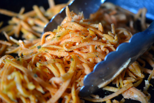 A pair of tongs is stirring the seasoned shredded sweet potatoes in the pan.