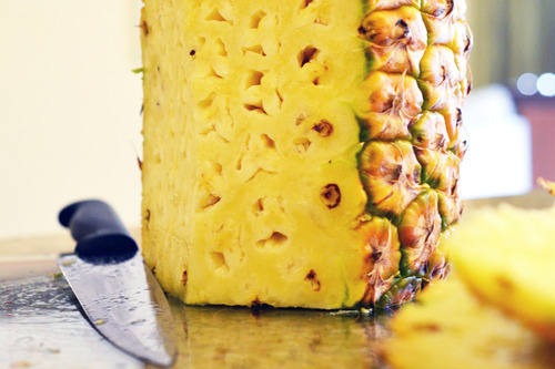 A half trimmed pineapple on a cutting board with a knife laying beside it.